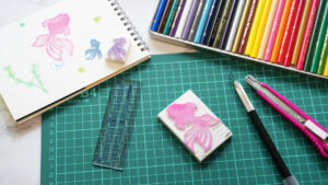 彫刻中の消しゴムはんことカッター、スタンプ道具が並ぶ制作風景。