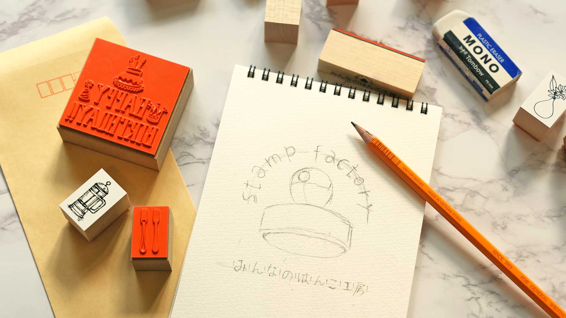 オリジナルスタンプとデザイン原稿、スタンプ台を並べ、完成イメージや作成ポイントを確認している様子を表現したイメージ画像。